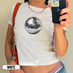 May include: A white short-sleeved t-shirt featuring a centered graphic of a silver disco ball with a grid pattern. The shirt is paired with a gold-toned chain necklace and an orange shoulder bag with a metal clasp and a small hanging charm.