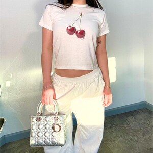 May include: A white cropped t-shirt with a graphic of two red cherries. The shirt is worn by a person with a silver handbag.
