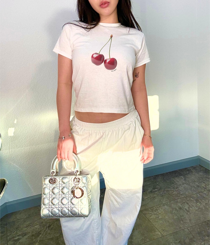 May include: A white cropped t-shirt with a graphic of two red cherries. The shirt is worn by a person with a silver handbag.