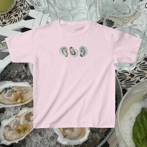 Pode incluir: Uma camiseta rosa com um gr&aacute;fico de tr&ecirc;s conchas de ostra na frente. As ostras est&atilde;o dispostas em fila, com a concha superior sobrepondo-se ligeiramente &agrave; concha do meio. As conchas s&atilde;o de cor castanho claro com um interior branco.