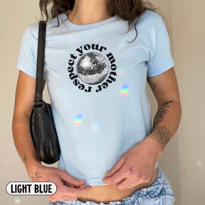 Puede incluir: Una camiseta de manga corta de color azul claro con un gr&aacute;fico de una bola de discoteca centrado en el pecho. El texto "respect your mother" est&aacute; impreso en una fuente negra circular alrededor de la bola. La camiseta tiene cuello redondo y corte ajustado.