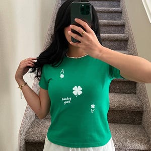 May include: Green short-sleeved t-shirt with a white four-leaf clover and the text "lucky you" printed on the front. The shirt also has two white ace of clubs playing card symbols.