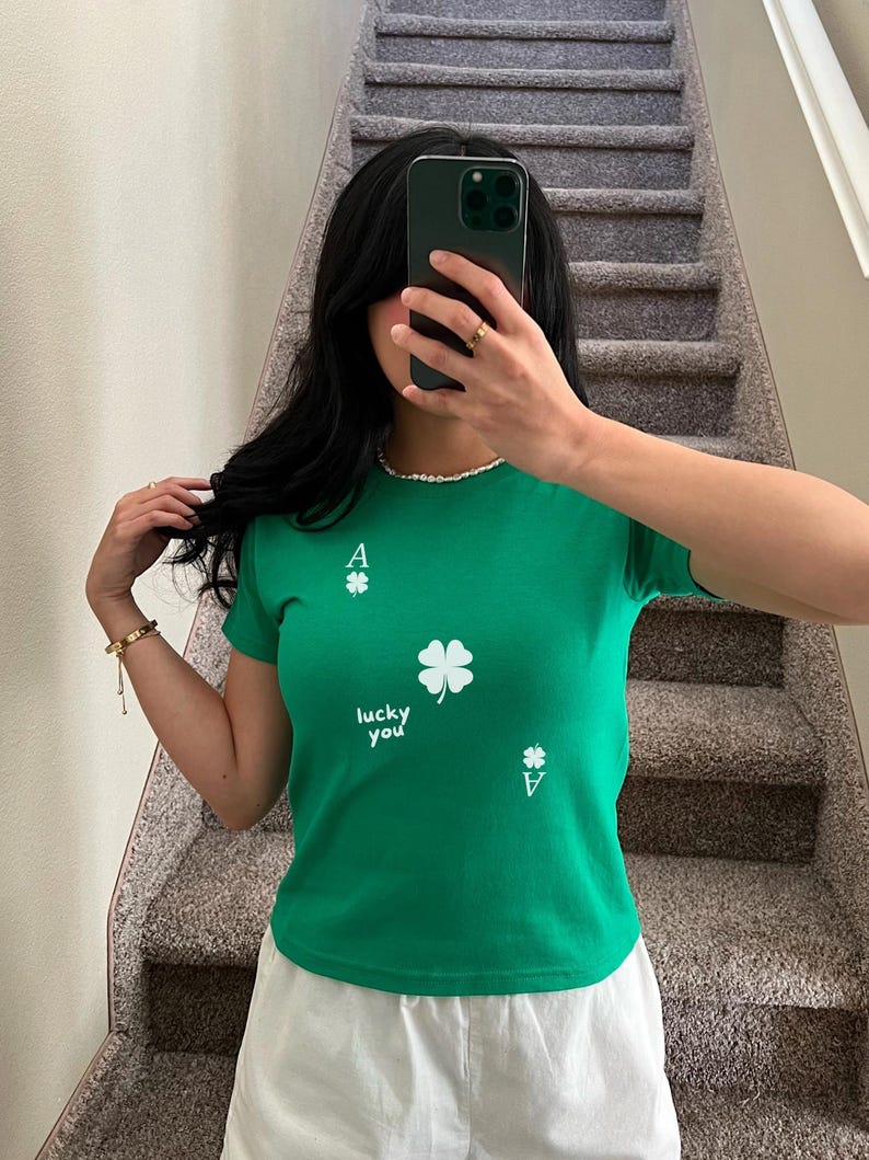 May include: Green short-sleeved t-shirt with a white four-leaf clover and the text "lucky you" printed on the front. The shirt also has two white ace of clubs playing card symbols.