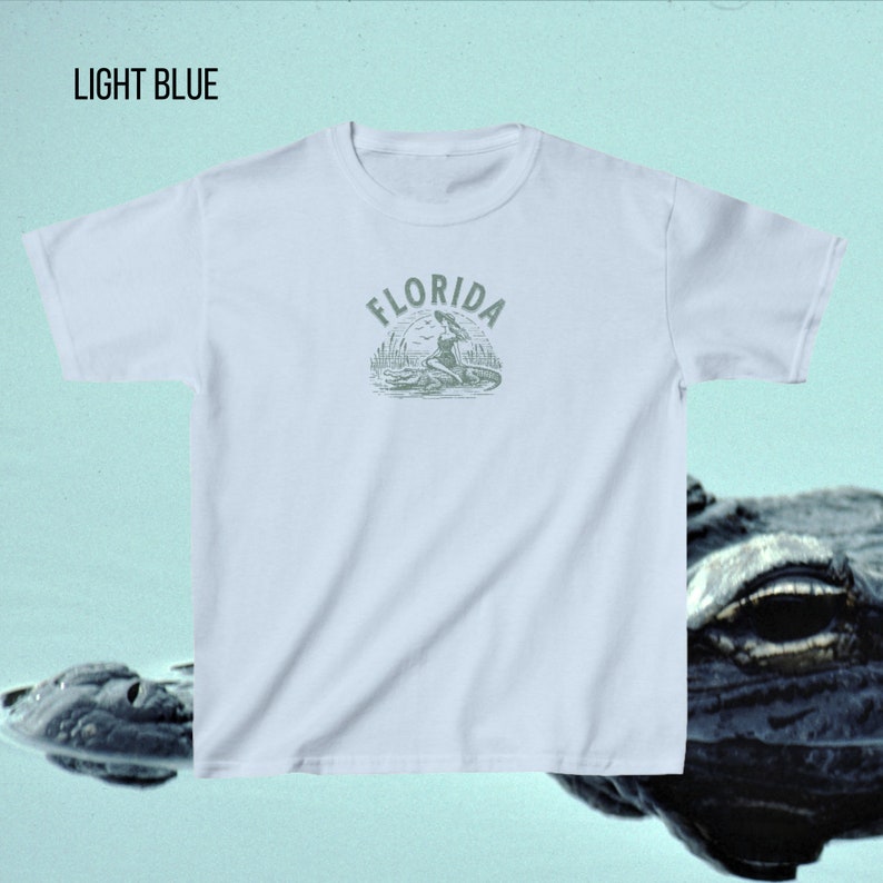 Puede incluir: Camiseta azul claro con la palabra "FLORIDA" encima de un gr&aacute;fico de una mujer sobre un caim&aacute;n. La camiseta es azul claro y tiene mangas cortas. El texto "LIGHT BLUE" est&aacute; en la esquina superior izquierda.