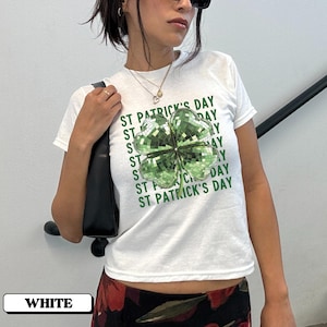 Puede incluir: Camiseta blanca con un diseño de trébol verde y el texto "St. Patrick's Day" repetido varias veces.