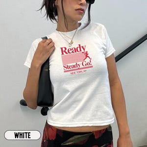 May include: A white short-sleeved t-shirt featuring a red graphic print on the chest. The design includes the text Ready Steady Go! with a silhouette of a running figure and horizontal lines, followed by the smaller text NEW YORK, NY below.