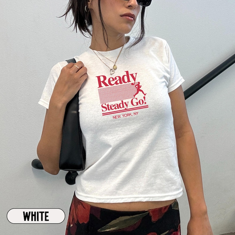 May include: A white short-sleeved t-shirt featuring a red graphic print on the chest. The design includes the text Ready Steady Go! with a silhouette of a running figure and horizontal lines, followed by the smaller text NEW YORK, NY below.