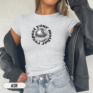 Puede incluir: Una camiseta gris claro de manga corta con un gr&aacute;fico central de una bola de discoteca plateada rodeada por el texto negro "respect your mother" en disposici&oacute;n circular. La camiseta se lleva bajo una chaqueta negra con vaqueros azules.
