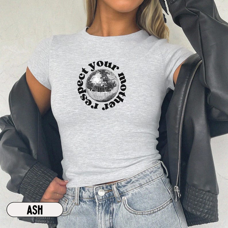 Puede incluir: Una camiseta gris claro de manga corta con un gr&aacute;fico central de una bola de discoteca plateada rodeada por el texto negro "respect your mother" en disposici&oacute;n circular. La camiseta se lleva bajo una chaqueta negra con vaqueros azules.