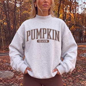 May include: A light gray heathered sweatshirt with a mock neck and long sleeves. The front features the text PUMPKIN in a large brown collegiate-style font, with the word SEASON printed in a smaller brown font inside a brown horizontal oval.