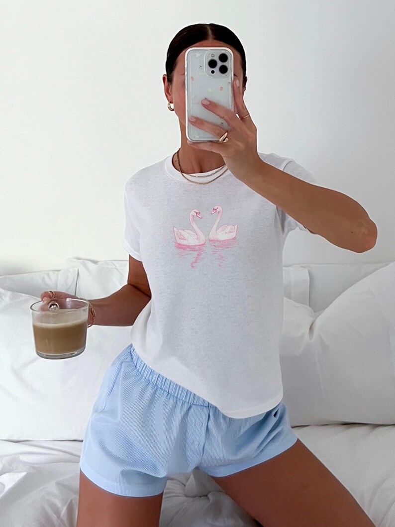 Puede incluir: Una camiseta blanca con un gr&aacute;fico de cisne rosa. La persona lleva pantalones cortos azul claro y sostiene una taza de caf&eacute;.