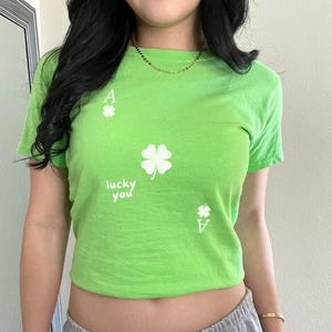 May include: A green cropped t-shirt with a white four-leaf clover and the text "lucky you" printed on the front. The shirt also has a white ace of spades printed on the front.