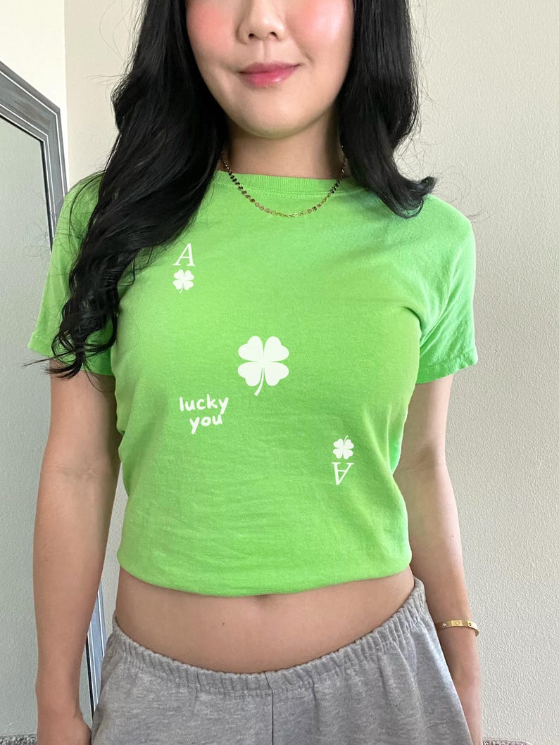 May include: A green cropped t-shirt with a white four-leaf clover and the text "lucky you" printed on the front. The shirt also has a white ace of spades printed on the front.
