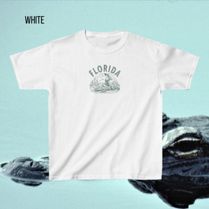 Puede incluir: Camiseta blanca con cuello redondo y mangas cortas. La camiseta presenta una ilustraci&oacute;n verde de un caim&aacute;n y una mujer con la palabra "FLORIDA" encima de la ilustraci&oacute;n. La palabra "WHITE" est&aacute; en la esquina superior izquierda.