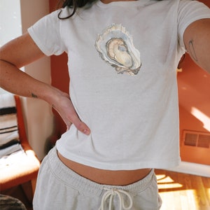 May include: A white t-shirt featuring a watercolour illustration of an oyster shell on the front.