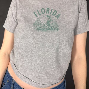 Puede incluir: Camiseta gris jaspeado con cuello redondo y mangas cortas. La camiseta presenta un gr&aacute;fico verde de la palabra "FLORIDA" sobre una ilustraci&oacute;n de una mujer, un caim&aacute;n y plantas de pantano.