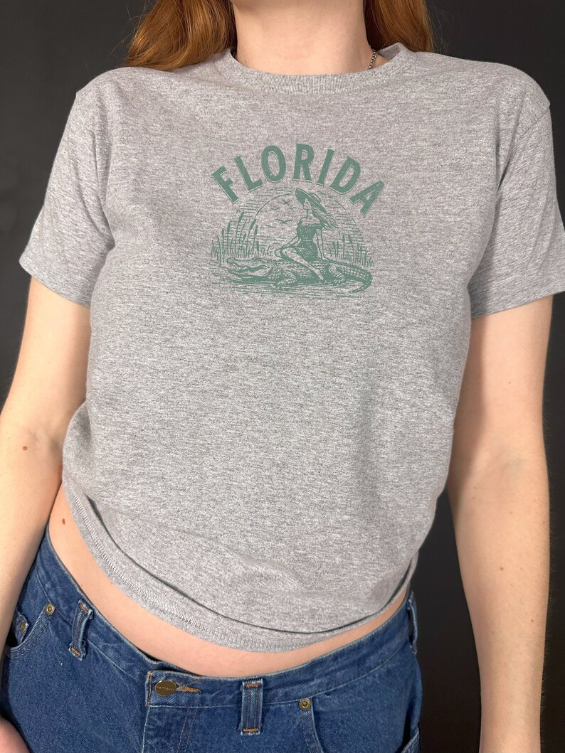 Puede incluir: Camiseta gris jaspeado con cuello redondo y mangas cortas. La camiseta presenta un gr&aacute;fico verde de la palabra "FLORIDA" sobre una ilustraci&oacute;n de una mujer, un caim&aacute;n y plantas de pantano.