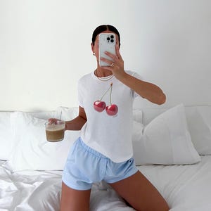 May include: A woman wearing a white t-shirt with a cherry graphic and light blue shorts. She is sitting on a white bed with a white pillow behind her. She is holding a cup of coffee in her left hand and a cell phone in her right hand.