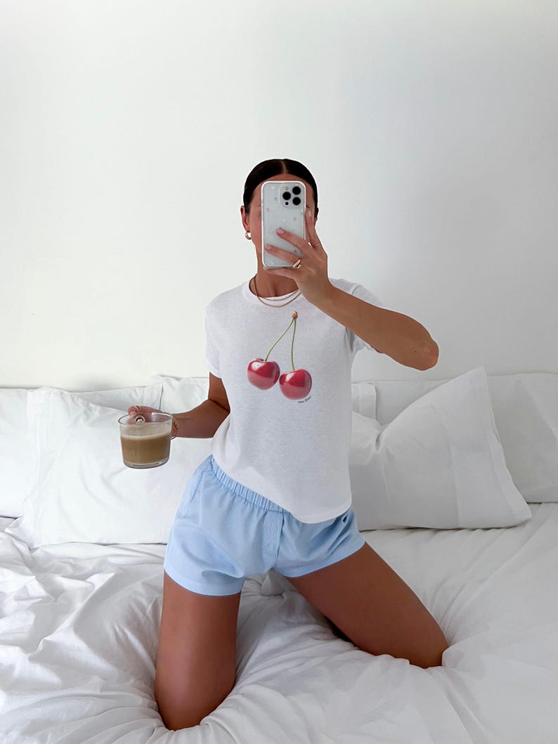 May include: A woman wearing a white t-shirt with a cherry graphic and light blue shorts. She is sitting on a white bed with a white pillow behind her. She is holding a cup of coffee in her left hand and a cell phone in her right hand.