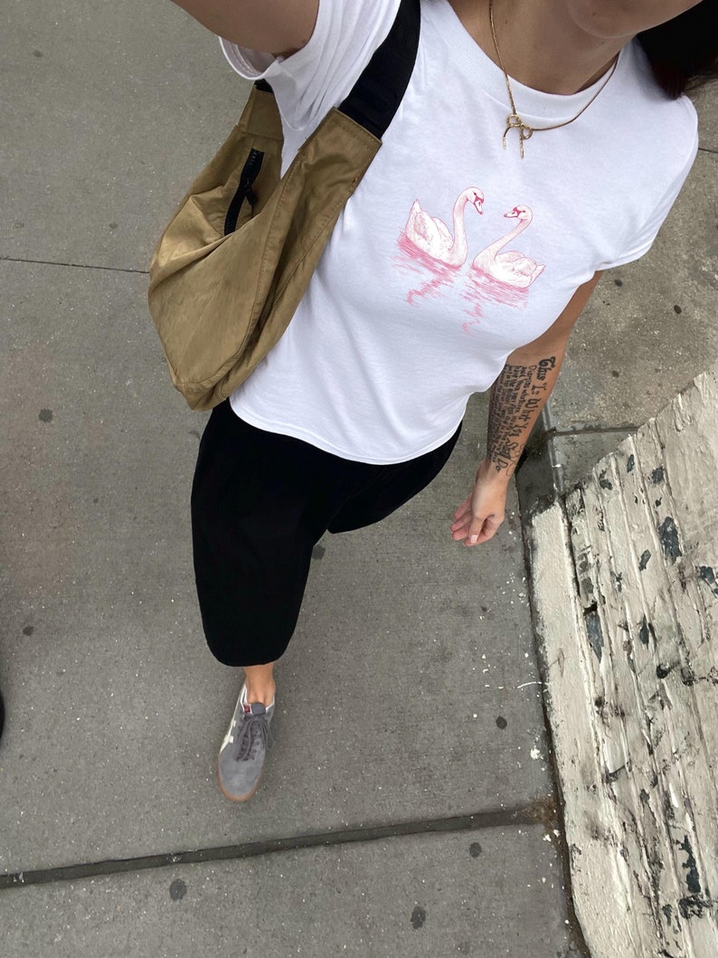 Puede incluir: Una camiseta blanca con un gr&aacute;fico de dos cisnes rosas en un lago. La persona lleva pantalones negros y una bolsa marr&oacute;n colgada del hombro.