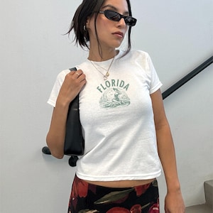 Puede incluir: Camiseta blanca con un gr&aacute;fico verde de una sirena y la palabra "FLORIDA". La camiseta se combina con una falda con estampado floral y gafas de sol negras. Un bolso negro se lleva al hombro.