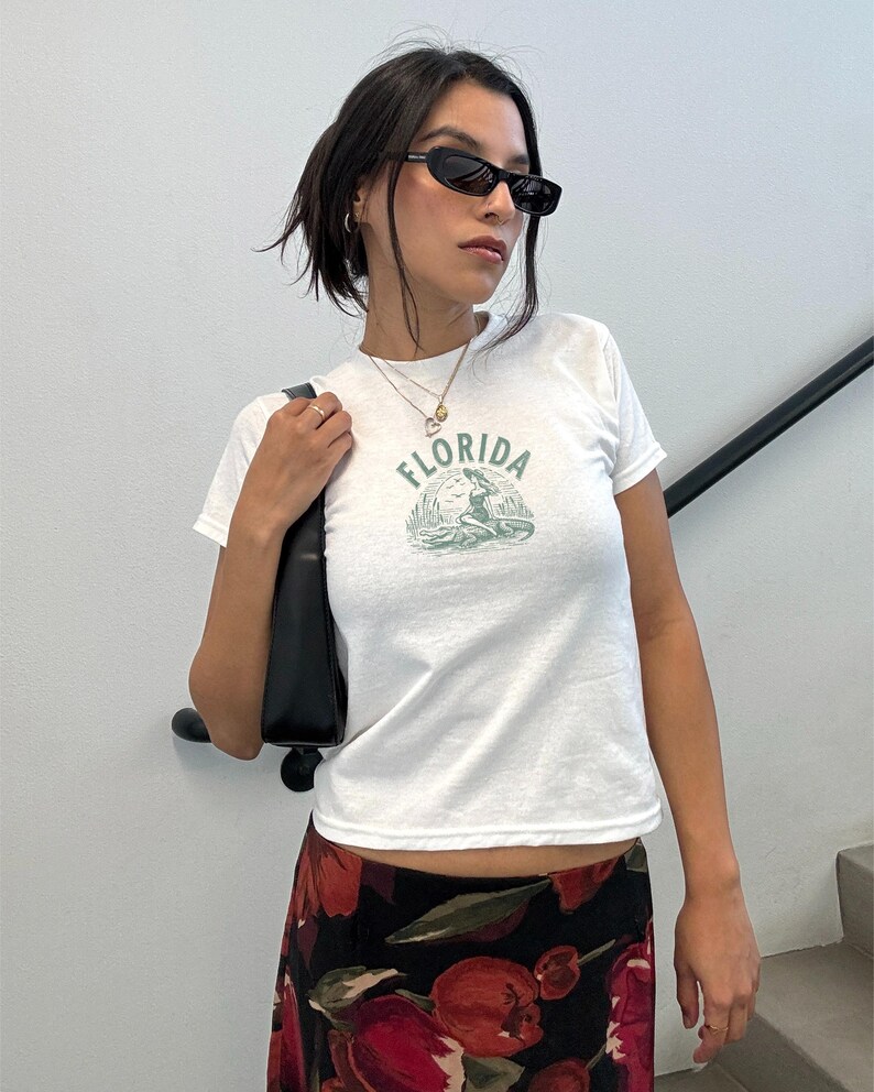 Puede incluir: Camiseta blanca con un gr&aacute;fico verde de una sirena y la palabra "FLORIDA". La camiseta se combina con una falda con estampado floral y gafas de sol negras. Un bolso negro se lleva al hombro.