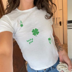 May include: White t-shirt with a green four-leaf clover and the text "lucky you" printed on it.
