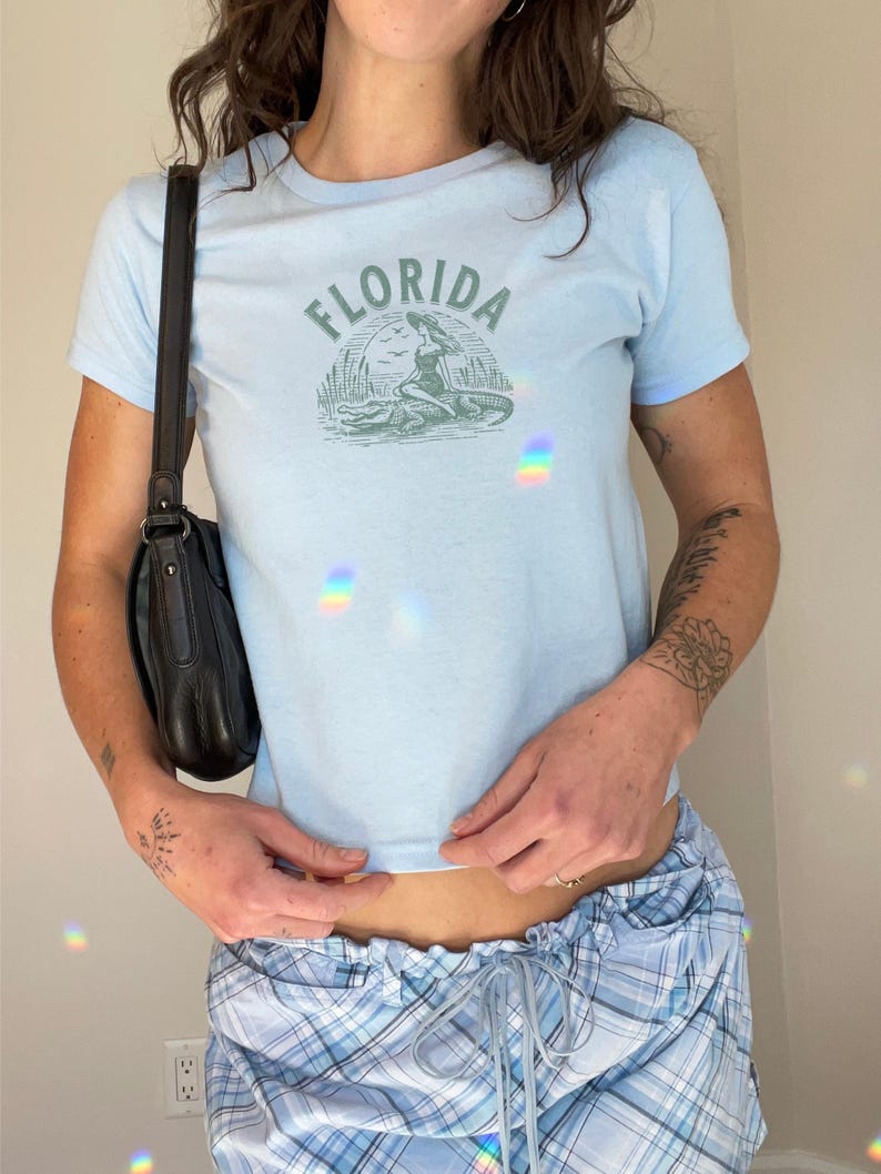 Puede incluir: Camiseta azul claro con la palabra "FLORIDA" y un gr&aacute;fico de una mujer sobre un caim&aacute;n. La camiseta se combina con pantalones cortos de cuadros azules. Se ve un bolso negro.
