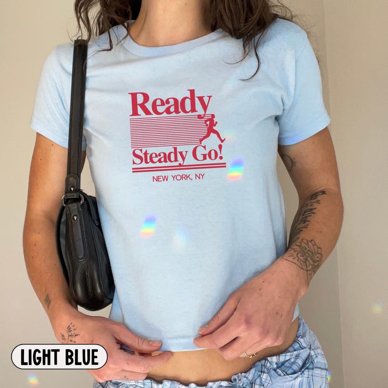 May include: A light blue short-sleeved t-shirt featuring red text that reads Ready Steady Go! with a graphic of a running figure and the words NEW YORK, NY. The shirt is paired with a black shoulder bag and blue plaid bottoms.