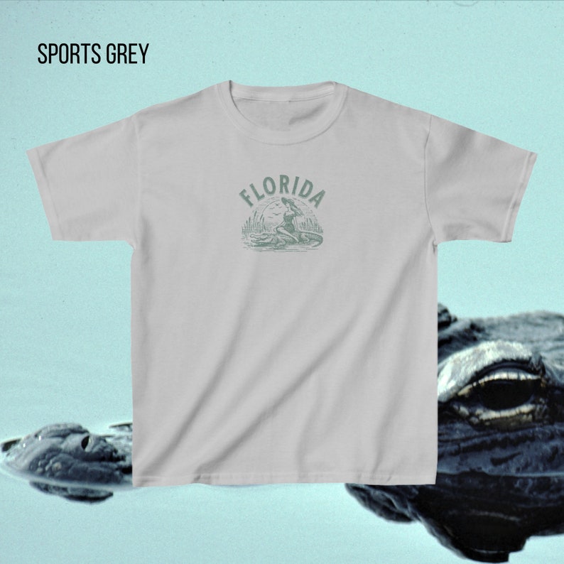 Puede incluir: Una camiseta gris claro con la palabra "FLORIDA" sobre un gr&aacute;fico de una mujer en un caim&aacute;n. La camiseta tambi&eacute;n tiene las palabras "SPORTS GREY" en la parte superior. El dise&ntilde;o es de color verde apagado.