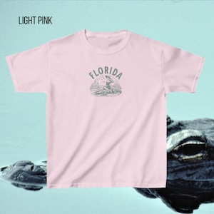 Puede incluir: Camiseta rosa claro con cuello redondo. La camiseta presenta un gr&aacute;fico de una mujer sentada en un caim&aacute;n con la palabra "FLORIDA" arriba. El texto "LIGHT PINK" est&aacute; en la esquina superior izquierda.