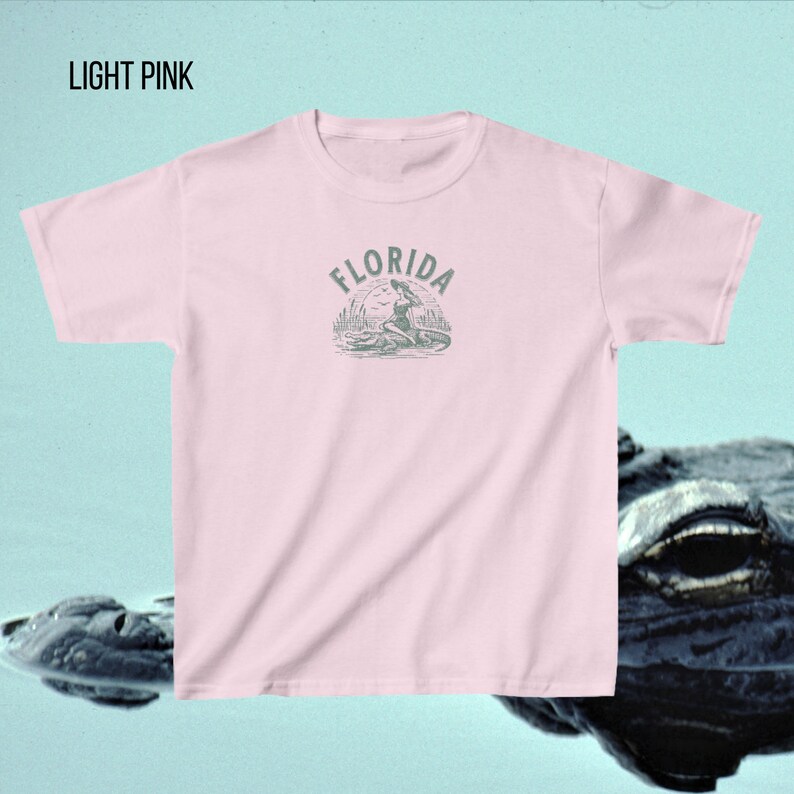 Puede incluir: Camiseta rosa claro con cuello redondo. La camiseta presenta un gr&aacute;fico de una mujer sentada en un caim&aacute;n con la palabra "FLORIDA" arriba. El texto "LIGHT PINK" est&aacute; en la esquina superior izquierda.