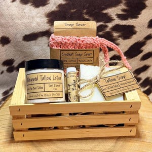 May include: A wooden crate filled with skincare products, including a jar of whipped tallow lotion, lip balm, and a bar of tallow soap. A pink crochet soap saver and a small wooden tag are also included. The products are displayed on a wooden surface.