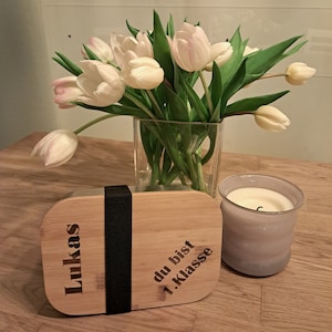 May include: A wooden lunchbox with a black elastic band. The lunchbox has the text "Lukas" and "du bist 1. Klasse" engraved on it. A bouquet of white tulips in a glass vase and a grey candle are on a wooden table.