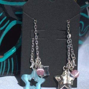 May include: A pair of silver dangle earrings with blue star charms and pink beads. The earrings are hanging on a black card.