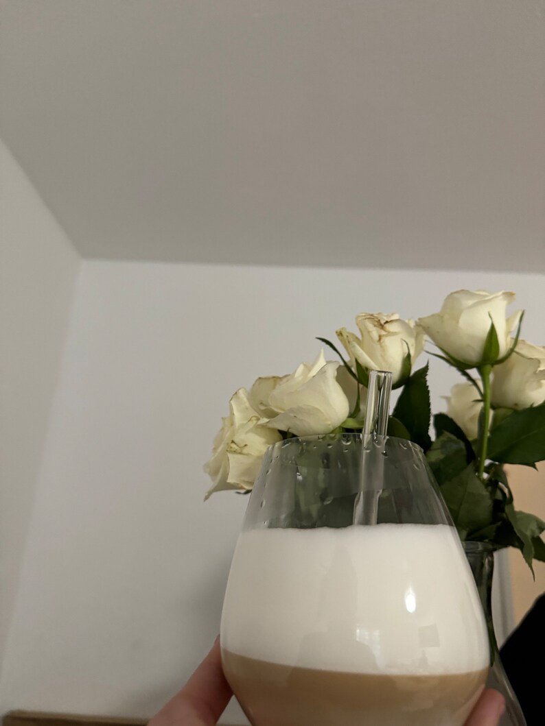 May include: A glass of coffee with a layer of foam and a clear glass straw, held in a hand. White roses are in the background.