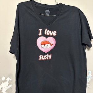 May include: Black v-neck t-shirt with a pink heart and the text "I love sushi" in brown. Inside the heart is a cartoon sushi roll with a smiling face.