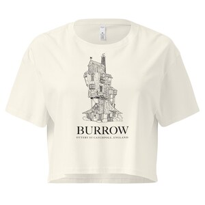 May include: Cream-colored, cropped t-shirt featuring a black and white illustration of a whimsical, multi-story house with the text "BURROW OTTERY ST CATCHPOLE, ENGLAND" below the image. The shirt has short sleeves and a crew neck.