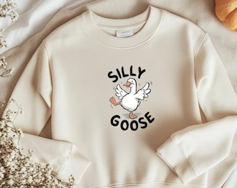 May include: A cream-coloured sweatshirt with a cartoon goose design. The goose is white with outstretched wings and the words "SILLY GOOSE" in black. The sweatshirt has a crew neck and long sleeves, suitable for casual wear.
