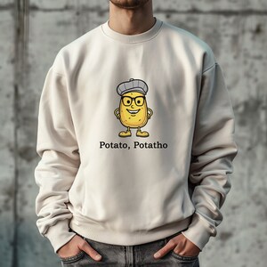 Pode incluir: Uma camisa de moletom de cor creme com um desenho de batata de desenho animado usando um chapéu e óculos. O texto "Potato, Potatho" é impresso abaixo da batata.