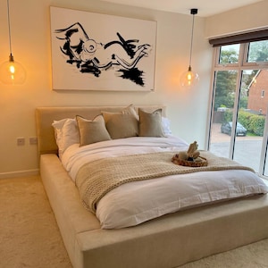 May include: A neutral-toned bedroom featuring a large upholstered bed with white bedding, and tan pillows. A black and white abstract artwork hangs above the bed. Two pendant lights with glass shades illuminate the space.
