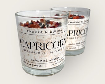 May include: Two glass candle jars with a white label featuring the zodiac sign Capricorn. The label includes the dates December 21 - January 19 and the text "CHAKRA ALQUIMIA". The candles are soy wax and have a scent of amber, musk, and nectarine. Each candle is 9 oz / 45 hours.
