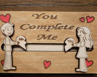 May include: Wooden plaque with the words "You Complete Me" engraved. The design features a man and woman holding puzzle pieces that fit together. Red hearts are scattered around the figures. A romantic gift.