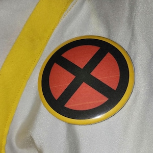 Op de afbeelding: Een ronde knoop met een gele rand, een rode kern en een zwart X-symbool. De knoop ligt op een witte stof met een gele streep.