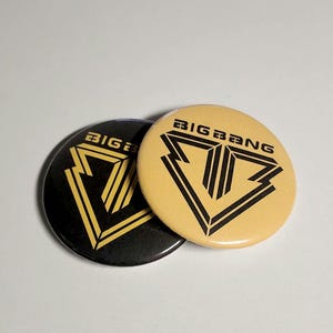 Pode incluir: Dois botões redondos com alfinete exibindo o texto BIGBANG e um logotipo geométrico em forma de V. Um botão é preto com um gráfico dourado e o outro é dourado com um gráfico preto. Os designs são idênticos e mostram um emblema estilizado.
