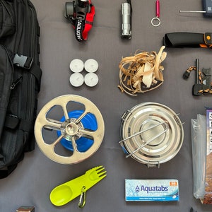 May include: A black backpack, a headlamp, a flashlight, a small pink keychain, a silver knife, a fire starter, a small metal pot, a camping stove, a set of three white tea lights, a pile of wood shavings, a green and yellow camping utensil, a pack of water purification tablets labeled "Aquatabs", a roll of duct tape, a small box of instant coffee, and a green tarp.