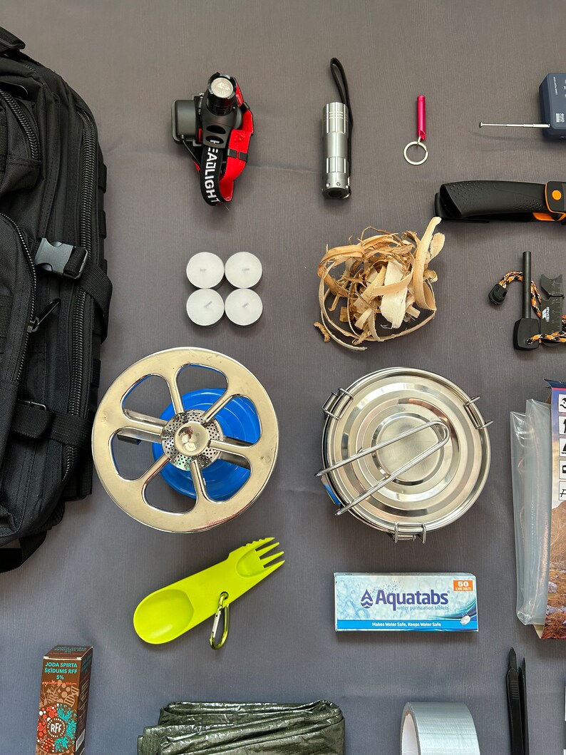 May include: A black backpack, a headlamp, a flashlight, a small pink keychain, a silver knife, a fire starter, a small metal pot, a camping stove, a set of three white tea lights, a pile of wood shavings, a green and yellow camping utensil, a pack of water purification tablets labeled "Aquatabs", a roll of duct tape, a small box of instant coffee, and a green tarp.