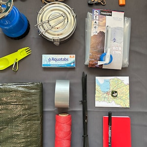 May include: A camping gear kit with a variety of items, including a blue camping stove, a silver food container, a green tarp, a roll of red string, a roll of gray duct tape, a water filter, a compass, a notebook, a pen, and a map.
