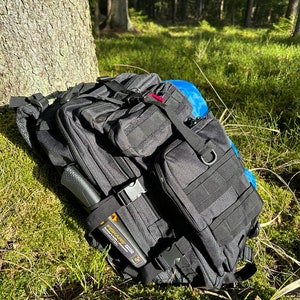 May include: A black tactical backpack with multiple pockets and straps, lying on a bed of green moss in a forest setting. A black knife with an orange handle is attached to the side of the backpack.