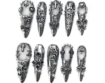 Puede incluir: Colección de diez anillos de uñas plateados con diseños góticos y religiosos. Cada anillo presenta cruces ornamentadas, flores de lis y motivos de corazón, algunos con pequeñas piedras transparentes. Los anillos se muestran sobre un fondo blanco.